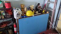 Wide shot showing the wood platform table with items on top including funnels, spray bottles, Roundup container, construction helmet, and various small tools and hardware.