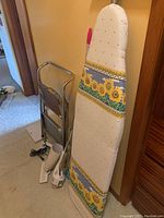 Photo of a sunflower patterned ironing board and metal step stool leaning against a wall next to an iron and handheld vacuum on the floor.