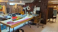 Wide view of crafting workspace featuring a large table with various crafting supplies including a magnifying work lamp and wood pieces, and a chair on wheels.