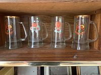 Four glass Oktoberfest beer steins with colorful cartoon designs and gold rims on a wooden shelf
