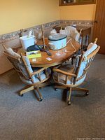 Oval wooden kitchen table with four matching rolling chairs upholstered with beige cushions, situated in a carpeted room with wallpaper border and window in background. The table has some items on it which are not included.