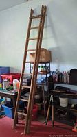 Wooden extension ladder leaned against wall beside metal shelving unit holding various items including pots, containers, and bottles.