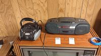 Two portable radios with CD players placed on a wood surface in front of a wood paneled wall, showing one Sony boombox and one smaller portable CD player radio.