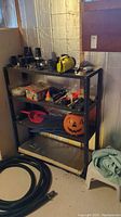 Black metal shelving unit filled with miscellaneous tools, containers, tubing on floor, and holiday decoration in basement setting.