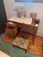 Wooden sewing cabinet with drawers open and floral cushioned stool placed in front. Sewing notions bag on top with a puzzle box.