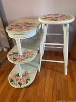 Three tiered wooden table and matching stool next to wall and mirror on floor with floral painted designs on white furniture.