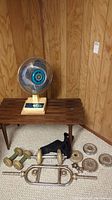 Lot showing oscillating fan on wooden slatted table with dumbbells, weights, trumpet, and case on floor.