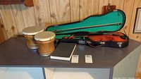 Violin in case with bow, two small bongo drums, and a hardcover book placed on a table.