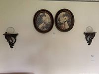 Wall display showing two oval framed vintage prints centered above paired carved wood wall sconces with clear decorative glass plates.