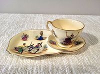 Side view of the tea cup placed on the uniquely shaped snack plate showing colorful painted dancers with gold trim on both items.