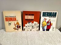 Photo showing front covers of three softcover books: The 1st Treasury of Herman (beige cover), The Second Herman Treasury (orange cover), and Herman The Sixth Treasury (white cover). Books placed side by side on a light fabric surface.