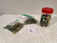 Three containers of vintage cats eye and solid marbles: two plastic resealable bags and one clear jar with red lid on a beige surface.
