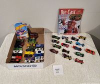 Overall photo of approximately 44 assorted Hot Wheels diecast toy cars arranged on a table with one car in original packaging and 2002 Die Cast Digest magazine in background.