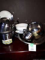 Stainless steel Shuttle Chef cooker and assorted pots with a white ceramic dish; cookware set on a dark wooden surface.