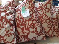 Set of four square cushions with a white floral pattern on a reddish background, shown stacked together and tagged with a lot number sticker.