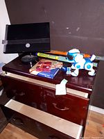 Wooden 2 drawer cabinet with various items on top including a black monitor, toy robot dog, colorful long toy sticks, and boxed games/cards.