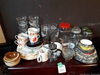 Wide view of all collected glassware items including glasses, mugs, plates, jars, and bowls together on a table.