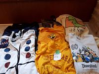 Photo of folded kids clothing including NASA hoodie, yellow shirts, dinosaur and Disneyland themed shirts.