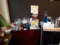 Wide view showing a collection of miscellaneous kitchen items on a dark wood dresser including dish racks, assorted plastic containers, metal and plastic utensils, ice packs, and various small kitchen ware items.