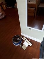 Photo showing a wall-mounted rectangular mirror leaning against a green wall, a round black-faced clock, a white multi-outlet power strip, a beige outlet adapter, and an extension cord on the floor.