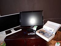 Two Samsung monitors on a wooden surface, one with white frame and one black SyncMaster T220, with power cables and a box containing Linksys router and accessories.