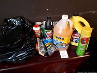 Photo showing various cleaning products and garbage bags on a dark surface including Drano, Comet, Old Dutch, Rain-X, gloves, and aerosol cans.