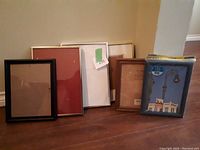 Six picture frames of various sizes and colors leaning against a wall, all with glass.