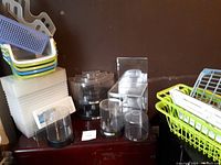 Multiple plastic holders including trays, clear display boxes and cylindrical holders, stackable plastic compartments, and small baskets. Items are stacked and arranged on a dark surface against a brown wall.