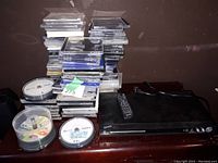 Overall view of the stack of rewritable CDs and DVDs with spindle cases and the CD/DVD player with remote on a wooden surface.