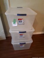 Three stacked Rubbermaid storage boxes with lids and blue handles on a wooden floor near a wall