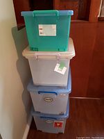 Stack of four plastic storage boxes in varying sizes and colors, showing lids and some brand stickers.