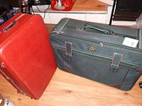 Photo shows two vintage suitcases from the 1960s, one red hard-sided and one green soft-sided, placed on a wooden floor.