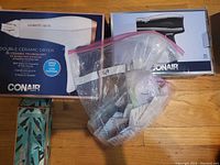 Two Conair hairdryers in packaging with clear plastic bag containing nail-less hooks on wooden floor
