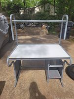 Front view of a bare desk with white surface, steel grey frame, and storage compartments underneath outside on driveway.