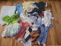 Full view of assorted baby and toddler clothing spread on a wooden floor, showing costumes, onesies, hats, and blankets.