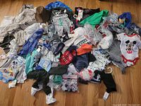 Photo showing assorted baby and toddler clothing including various onesies, rompers, hats, and socks spread out on wooden floor