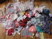 Lot of assorted baby, toddler and kids clothing spread out on floor. Multiple tops, dresses, leggings, and onesies in mixed colors and patterns visible.