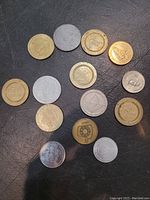 Overview of 15 gold and silver metal casino tokens and chips with various logos and numbers, laid out on black surface.