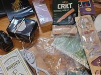 Top-down photo showing a variety of miscellaneous items including a CRKT knife kit, Bossies clips, and a sealed pack of wooden and paper goods.
