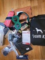 Overview of the lot showing black Team K9 tote bag, green and black Happy Dog harness, packaged choke collar, various boxed and wrapped items, balls, and electronics accessories.