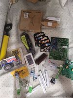 Photo showing assorted miscellaneous items including a yellow handled tool, boxed monitor stand, cables, circuit boards, and collectibles.