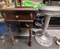 Image showing a dark carved wooden nightstand with turned legs and a drawer beside a metal and grey cushioned adjustable barstool.