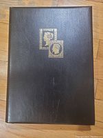 Photo showing front cover of a black deluxe stamp album with gold-embossed vintage British stamp designs