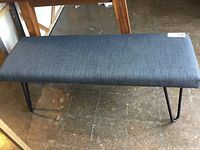 Front view of rectangular upholstered bench with blue-gray fabric upholstery and black metal hairpin legs on a concrete floor.