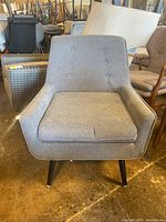 Front view of a gray upholstered lounge chair with button tufting and black angled legs on a concrete floor.