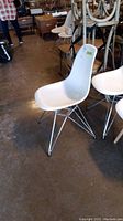Side view of a white molded plastic chair with chrome legs and crisscross supports, showing the full chair including backrest, seat, and legs on a concrete floor in a warehouse-like setting.