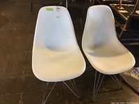 Pair of white plastic chairs with metal wire legs seen side by side on concrete floor.