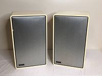 Front view of pair of vintage Advent speakers with metal grille covers, showing beige cabinets and black perforated metal front panels.