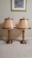 Two full view brass lamps with beige lampshades and black trim, on carpeted floor against a wall.