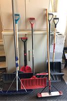 Full view of the lot showing five shovels and one push broom with handles, standing upright against a cabinet.